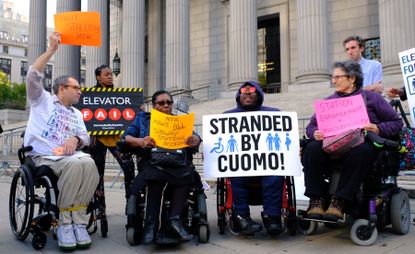 DIA members and other activists are outside of a building in downtown Manhattan protesting for the right to add elevators for all public transit stops. They are holding signs that say "Stranded by Cuomo" and "Elevator FAIL."