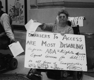 A black and white photo of a woman in a wheelchair holding a sign with black texts that reads: "Barriers to access are most disabling. Enforce ADA = Rights. Equal Access to all forms of transportation" with drawings of taxis, buses, ferries, and airplanes at the bottom.
