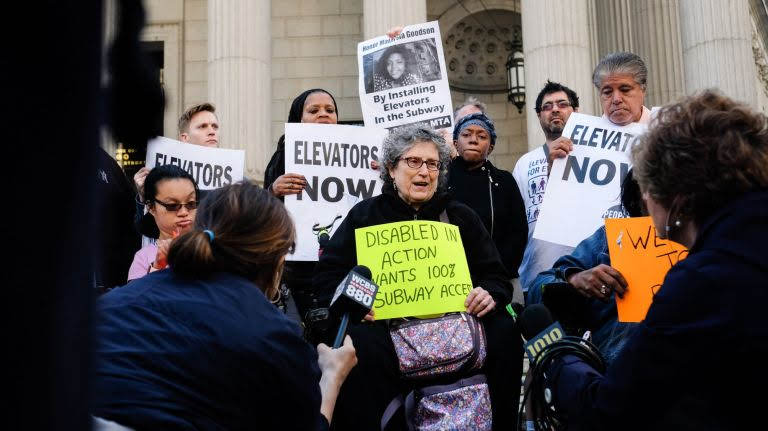 DIA members and other activists protesting for NYC subway accessibility in front of a courthouse, with news reporters holding microphones towards them. One woman is holding a sign that reads “Disabled in Action wants 100% subway access.” Behind her are people holding signs that say, “Elevators NOW.”