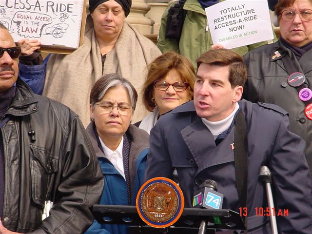 A male Access-A-Ride user, with dark brown hair and in a navy blue trench coat, talks about unsafe vans at a press conference.