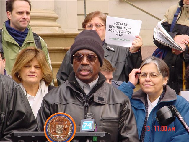 A gentleman wearing a black knit hat, dark sunglasses, a black leather jacket and a white collared shirt speaks at the podium in front of City Hall.