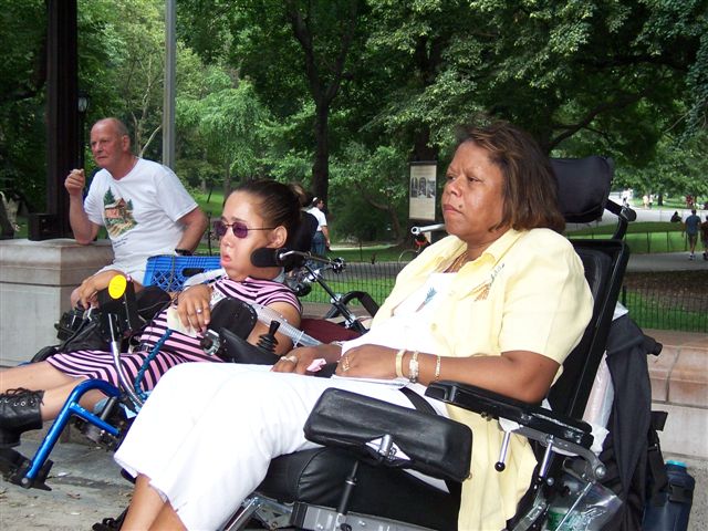A couple of disabled people in wheelchairs are seen listening to speeches in the park