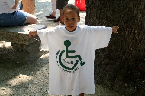 A boy, standing with outstretched arms, wears a T-shirt that bears the logo of ICS(Independence Care System), which includes the wheelchair sign.