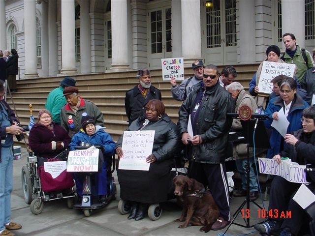 Outside of City Hall on March 13, 2003, a group of people, some standing and some sitting in wheelchairs and scooters, are surround Councilwoman Margarita Lopez holding a piece of paper. Most of the people in the group are holding up signs such as "REVAMP ACCESS-A-RIDE NOW", "CHILDREN CAN TOO RIDE ACCESS-A-RIDE!", "DISABLED IN ACTION", and "Bad Service! Access-A-Ride Stinks! Riders Won't Take It Anymore".