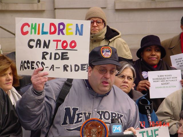 Some people surround a man wearing glasses, a dark blue baseball cap bearing the red and orange Fire Department of New York City logo, and a light gray snap-button jacket reading "NEW YORK" in block letters with a wavy line underneath the words "NEW YORK". The man holds up a sign reading "CHILDREN CAN TOO RIDE ACCESS-A-RIDE!" while speaking behind the podium holding the New York City government logo in an orange and purple circle and a microphone representing Crosswalks Television Channel 74. Behind the man is Councilwoman Margarita Lopez wearing a blue parka. Behind Lopez is a woman, wearing a black hat and coat, holding up a sign reading "9,000 RIDES SCHEDULED BUT HOW MANY ARE ACTUALLY GIVEN? DISABLED IN ACTION"