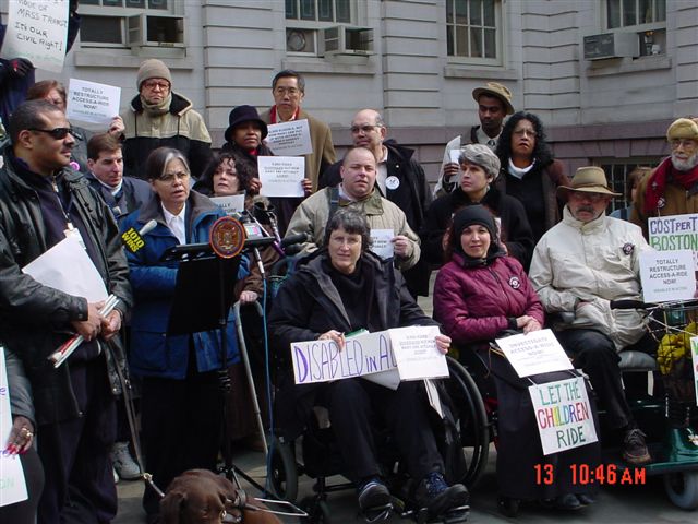Councilwoman Margarita Lopez is seen speaking at the press conference in front of City Hall surrounded by fellow protesters standing as well as in wheelchairs while most are holding up various signs protesting Access-A-Ride.