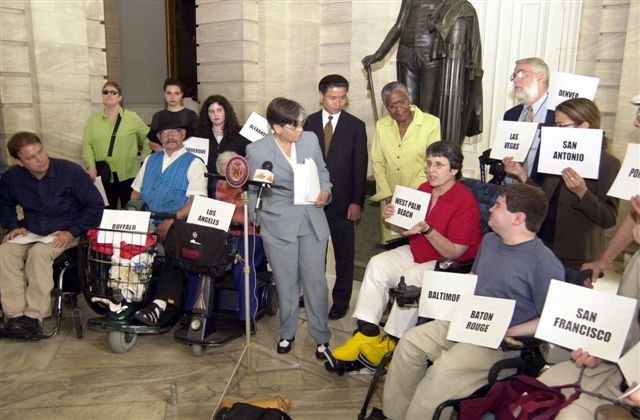 A group of about 14 people, some in wheelchairs and scooters, are standing around Councilwoman Margarita Lopez at a podium that holds one microphone. Lopez looks to her left side (our right side). Most of the people in the group are holding up pieces of paper stating names of cities such as "BUFFALO", "LOS ANGELES", "WEST PALM BEACH", "BALTIMORE", "LAS VEGAS", "BATON ROUGE", "DENVER", "SAN FRANCISCO", and "SAN ANTONIO".