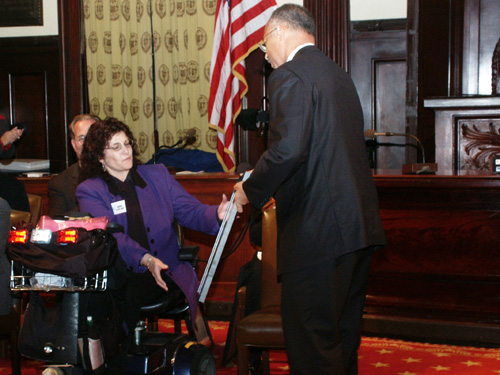 Inside a room at City Hall, Anita Apt, sitting in her scooter, receives an award which is seen being handed to her from New York City Comptroller William J. Thompson Jr. The ceremony was held on October 27, 2003 in recognition of achievement for disability awareness.