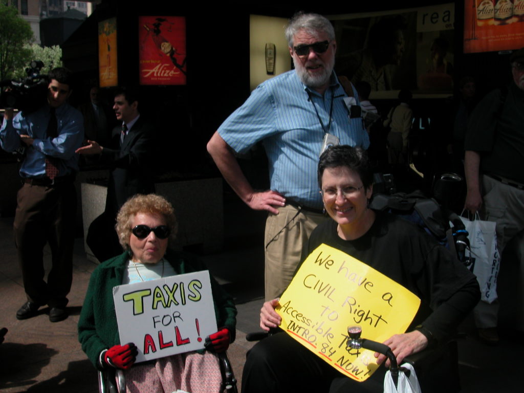 Two women in wheelchairs hold up signs reading "TAXIS FOR ALL!" and (on a yellow cardboard) "WE HAVE A CIVIL RIGHT TO ACCESSIBLE TAXIS - INTRO. 84 NOW!". Behind the two women stands a man with a beard in blue shirt and sunglasses. To the far left, in the background, are two men, one of which holding a portable television camera.
