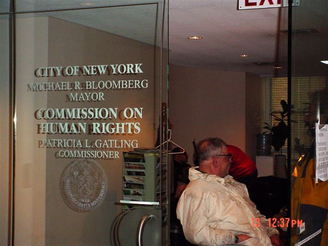 The front glass door of the Human Rights Commission office with white text on the glass reading "CITY OF NEW YORK MICHAEL R. BLOOMBERG MAYOR COMMISSION ON HUMAN RIGHTS PATRICIA L. GATLING COMMISSIONER" with the New York City government logo traced in white underneath the words. At the front door is a male senior citizen in a white parka sitting in his wheelchair and looking in the office.