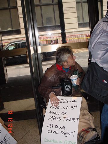 A woman in a dark brown leather jacket sitting in a wheelchair in the lobby of the Human Rights Commission looks to her left (our right) and holds a blue and white plastic cup full of coffee in her left hand while holding a sign in her right hand that reads "ACCESS-A-RIDE IS A THIRD MODE OF MASS TRANSIT. IT'S OUR CIVIL RIGHT! DISABLED IN ACTION" Her right hand covers the word "ACCESS" on the sign.