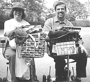 Ellen, wearing a baseball cap, is to the left of Jerry. Both are sitting in their own scooters and are situated in a park. Both of their baskets that come attached with the scooters are filled with clothes and bags.