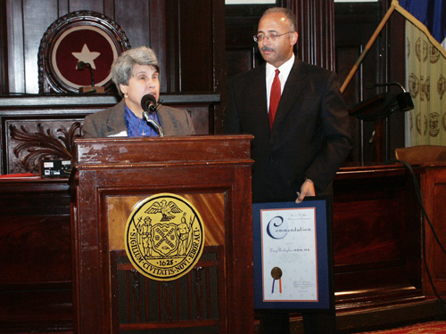 Inside a room at City Hall and standing at a podium bearing the New York State logo, Lucy Birbiglia speaks after receiving an award from the person standing to the right (her left), New York City Comptroller William J. Thompson Jr., who is seen holding the award. The ceremony was held on October 27, 2003 in recognition of achievement for disability awareness.