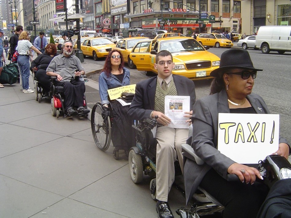 On the sidewalk in front of Manhattan's Penn Station, five people in wheelchairs line up for their so-called "chance" at a taxi. The woman in front of the line, in a motorized wheelchair and wearing a hat and sunglasses, holds a sign reading "TAXI!", while two other people in wheelchairs behind the woman holds signs with letters too small to read from this picture view.