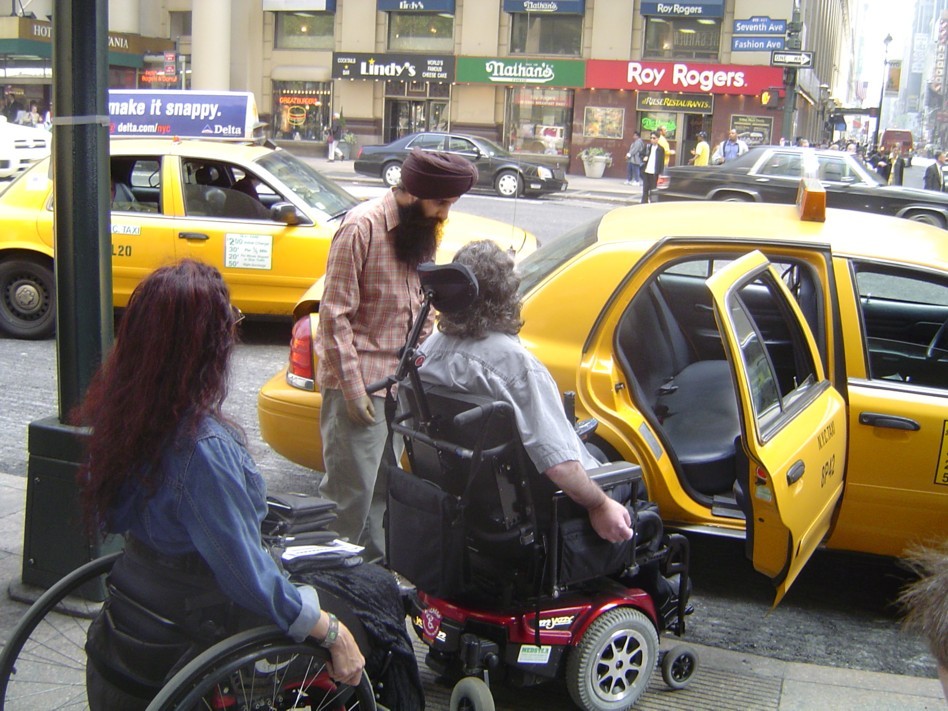 In front of Manhattan's Penn Station, a man in a motorized wheelchair with a red base is on the sidewalk and parked in front of an open door of a small yellow cab car. Behind the motorized-wheelchaired man is a woman in a blue denim, long-sleeved shirt and using a manual wheelchair while in front of the motorized-wheelchaired man is the cab driver looking at the open car door.
