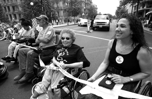 Carr and Nadina LaSpina at the May ADAPT Action. They, along with other protesters, are in wheelchairs in the street with white tape on each of their laps.