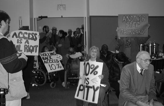 This photo was taken from inside a meeting room looking toward the door. Blocking the doorway are 2 people in power wheelchairs. The woman is carrying a sign that says, "Rolling Back Access ten percent" and the sign also has a smiley face on it. In the foreground on the right side, another woman is facing the photographer with a sign that says, "Ten percent is Pity." Four other people of various ages and races are near her. One man is wearing a tie, and the others are casually dressed. One person is holding up a sign that says, "Another step backwards." That person is wearing an ADAPT T-shirt. Someone on the left has a sign that says, "Access is a Civil Right." Although the doorway is wide, no one can pass through it because of the wheelchairs. Just on the other side of the two people blocking the door with their chairs are a man and a woman talking. The man is wearing an ADAPT T-shirt. In back of them are 3 State Troopers and a man in a suit who are looking into the room. Another man in a suit is looking in another direction.
