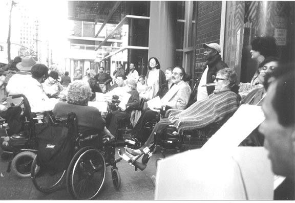 Twenty adults of different ages and races, most in wheelchairs and scooters, but some also standing, are facing each other in a layered circle outside a modern building. Some of them have signs. In the background, there is a bored-looking policeman with his arm across his chest. The people look like they are waiting for something.