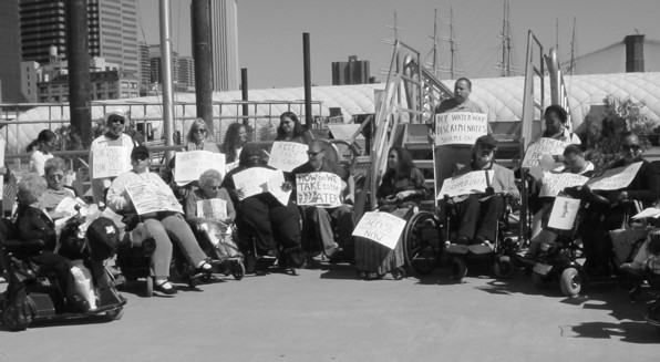 A group of about 20 wheelchair users and standees is demonstrating next to a set of ferry steps.