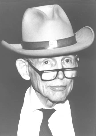 This picture of Justin Dart shows him as a senior citizen looking right into the camera with a steady gaze. He is wearing a big cowboy hat and his glasses are sliding down his nose. He is also wearing a white shirt and tie.