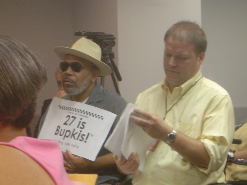 Photo of man, with Alexander Wood sitting next to him, holding sign reading "27 is bupkis"