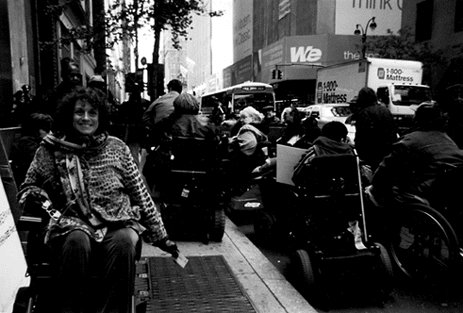 Simi Linton, a disability rights consultant, author, and teacher, is facing us and smiling. She uses a wheelchair. In the background are demonstrators at the Access-A-Ride demonstration on April 30, 2002, at MTA headquarters.