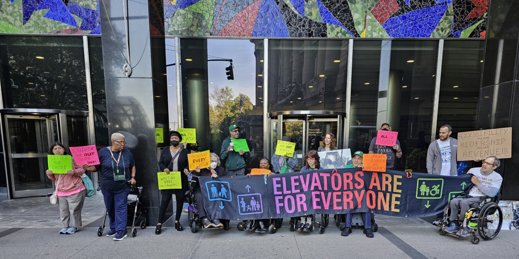 A group of demonstrators hold signs including "Elevators Are For Everyone," "Accessibility means Ridership means Revenue," "Maintain ALL the Elevators," 30 Elevators Are OUT Every Day," "A Broken Elevator Is an Emergency," "MTA: Quit Wasting $$$$ on Lawyers" and "Settle the Case"